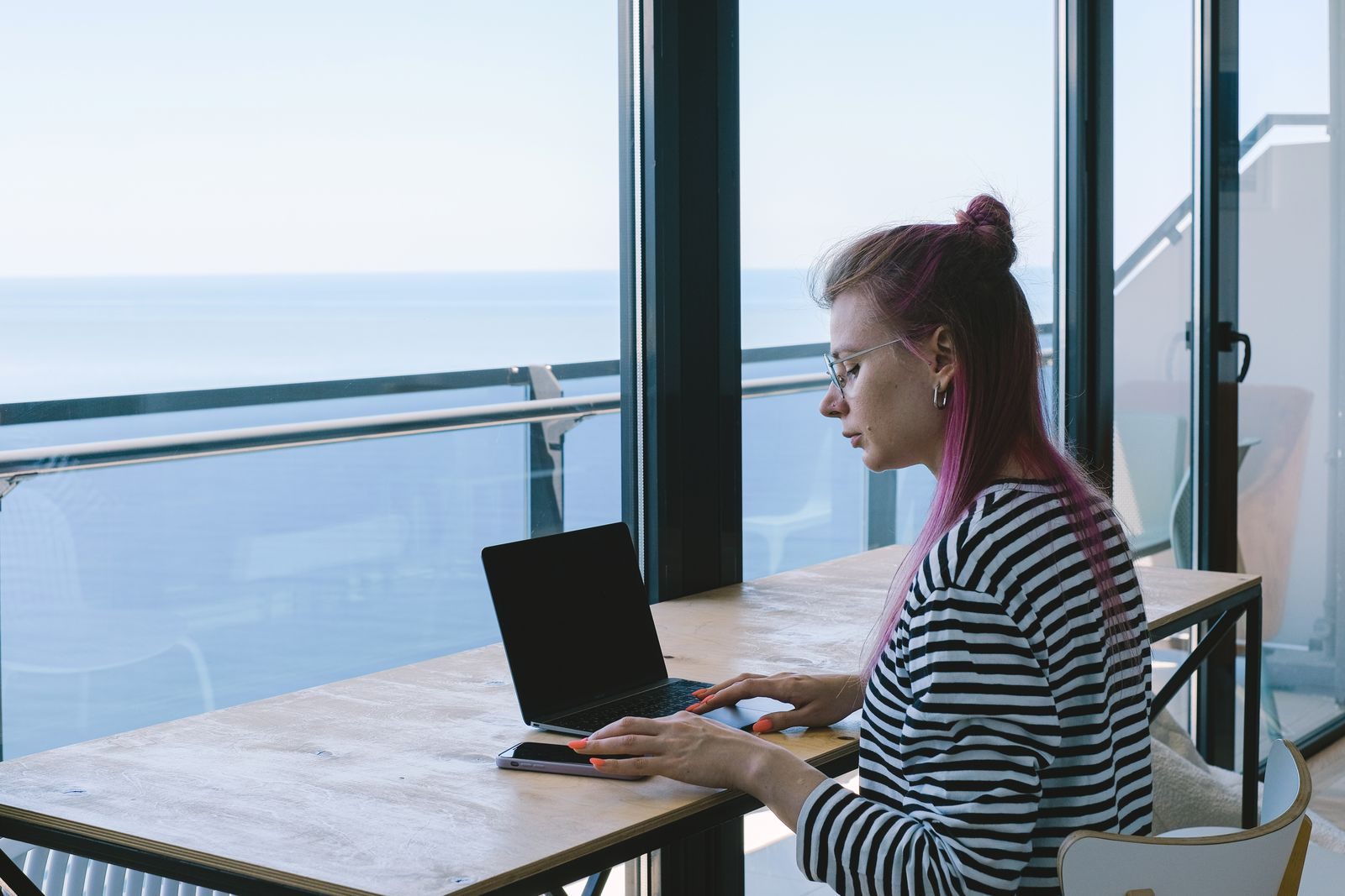 Woman working on a laptop at a table beside a window overlooking the sea